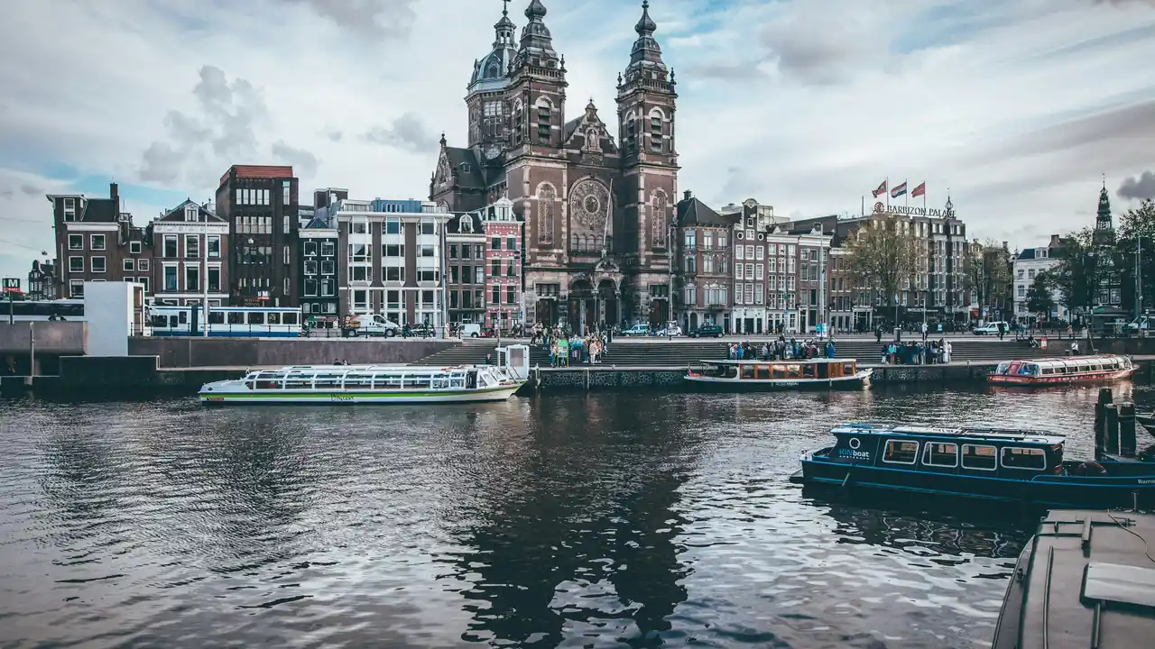 Amsterdam canal view with boats in the foreground and the Basilica of St. Nicholas near Amsterdam Central Station, showcasing historic architecture and waterfront cityscape in the Netherlands.