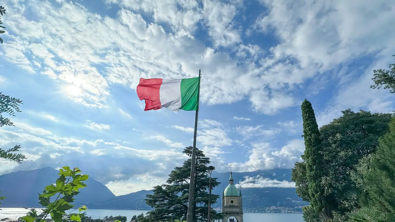 Italian flag waving on flagpole above scenic Lake Como with mountains, church dome, and dramatic cloudy sky in Italy