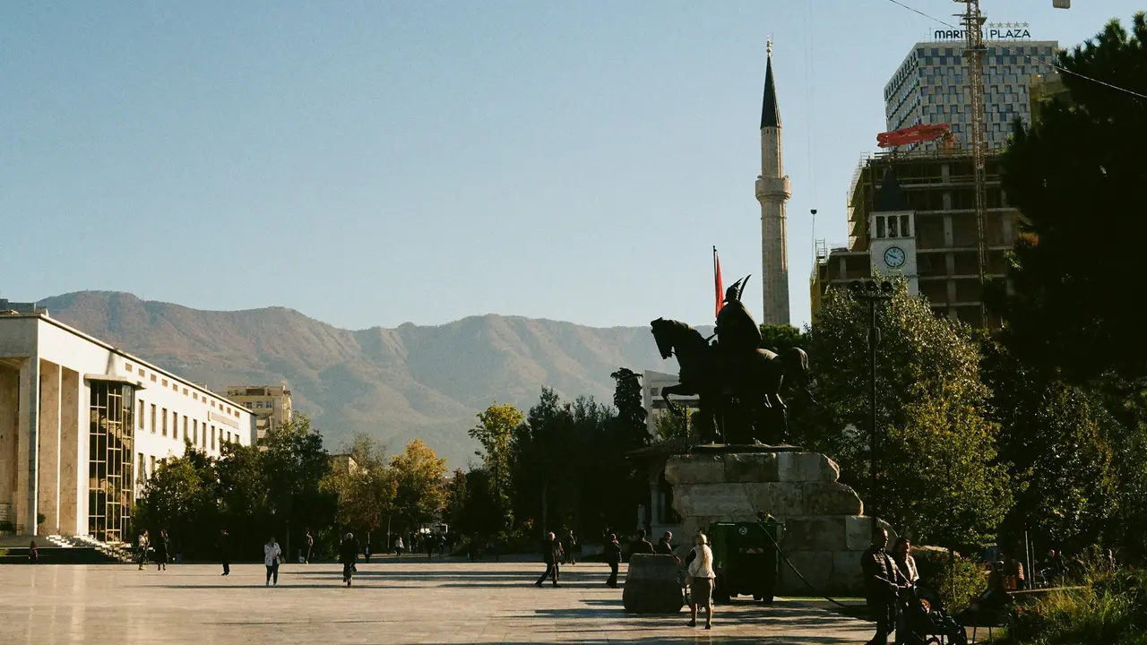 City square with equestrian statue and mosque minaret against mountain backdrop with modern buildings and pedestrians in urban landscape