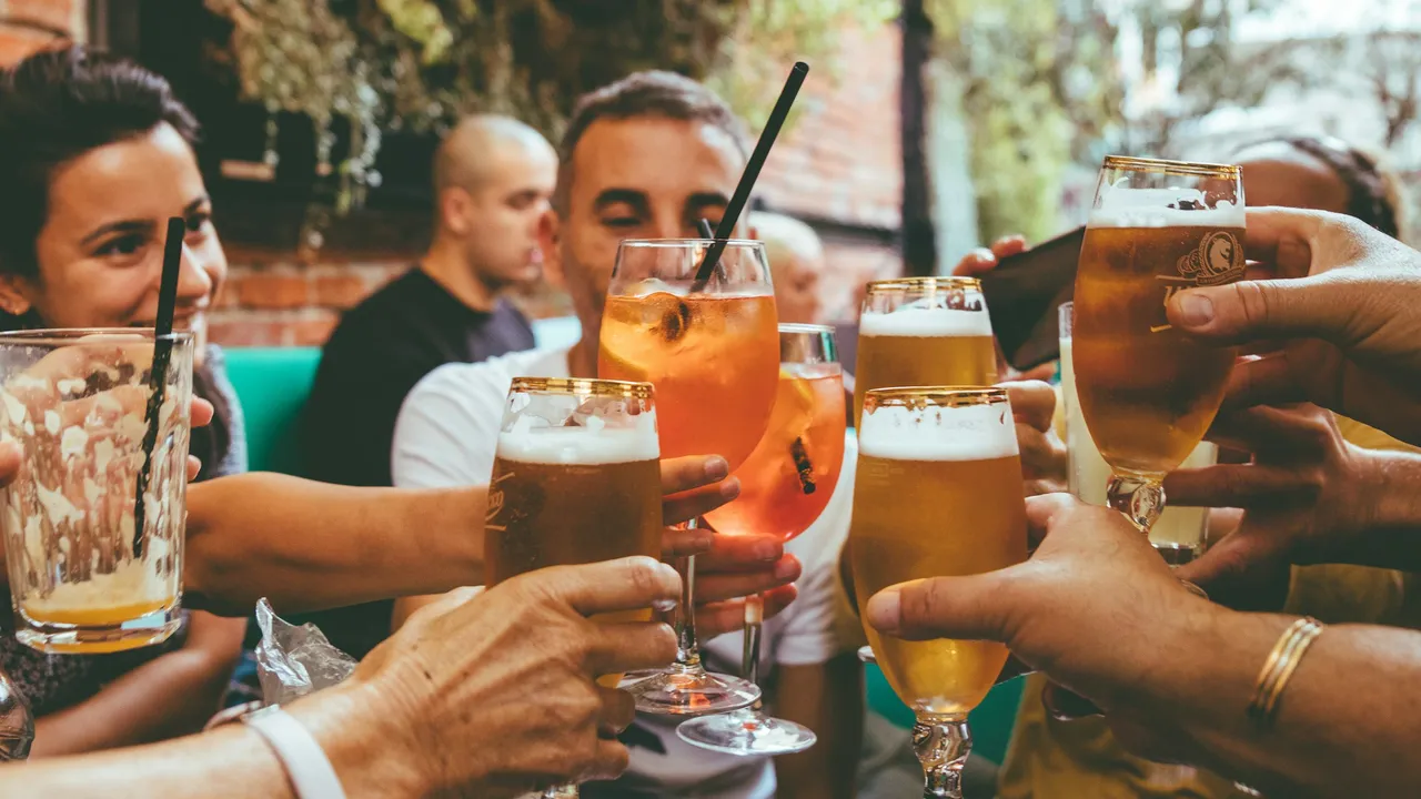 Friends clinking glasses of beer and cocktails at an outdoor bar, celebrating together in a lively social gathering during daytime.