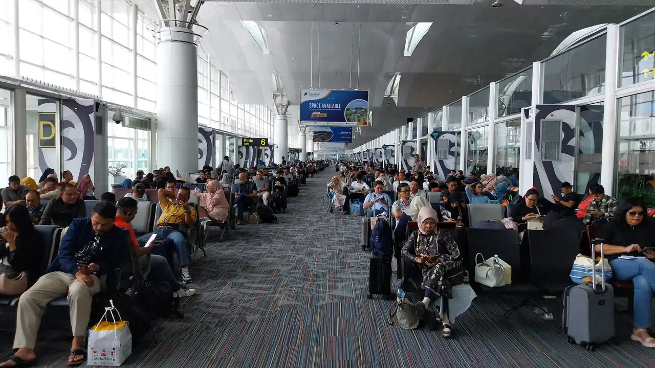 Busy airport departure lounge with passengers seated in long rows, waiting at boarding gates with carry-on luggage inside a modern international terminal.
