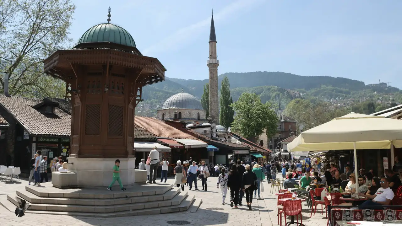 Sebilj Fountain in Baščaršija, Sarajevo old town, with traditional market streets, mosque minaret, cafes, and people walking on a sunny day