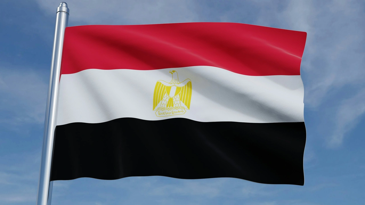 The Egyptian national flag waves against a bright blue sky with light, wispy clouds. The flag features three horizontal stripes of red, white, and black, with the golden Eagle of Saladin centered in the middle white stripe. The flag is attached to a silver metal pole on the left side of the frame.