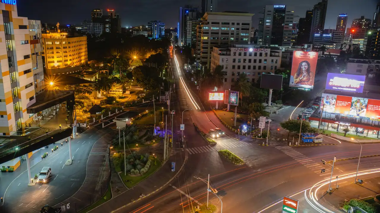 Long-exposure light trails from passing cars stretch across the asphalt, framed by multi-story buildings, glowing billboards, and lush tropical greenery. The modern architecture and vibrant street lights create a busy, urban atmosphere against the dark night sky.