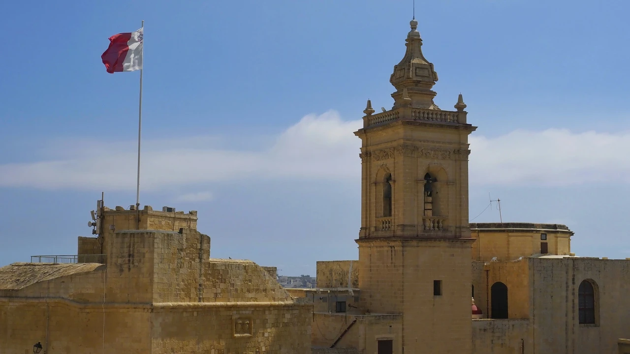 A close-up shot of a white stone bell tower in Malta, decorated for a local celebration. The tower features a classical design with arched openings and a large clock face. A Maltese flag, characterized by its red and white vertical stripes with the George Cross in the upper corner, hangs prominently from the tower. The bright red and white of the flag contrast against the weathered limestone of the architecture and the clear blue sky in the background