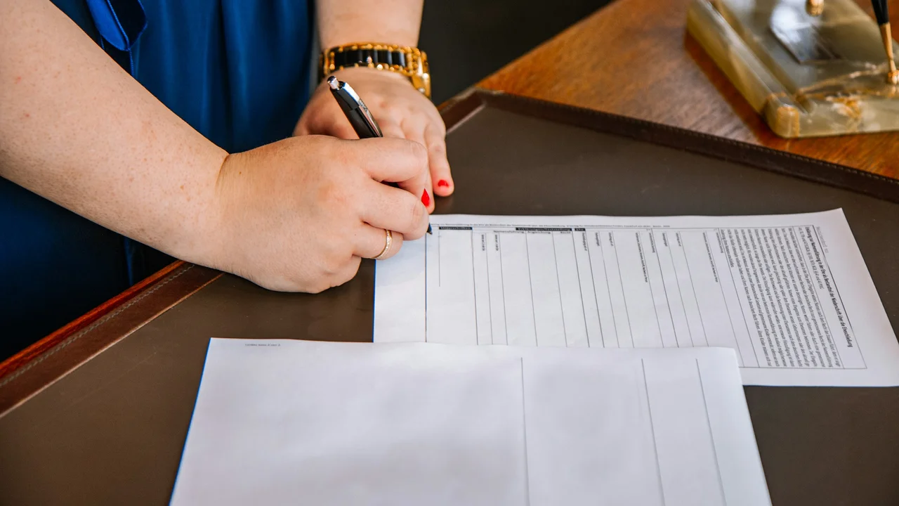A close-up shot of a person's hands as they sign a formal document on a dark desk mat. The individual is wearing a gold watch and ring, using a black pen to fill out a paper that appears to be a German visa or legal application. In the foreground, another partially visible document rests on the desk.