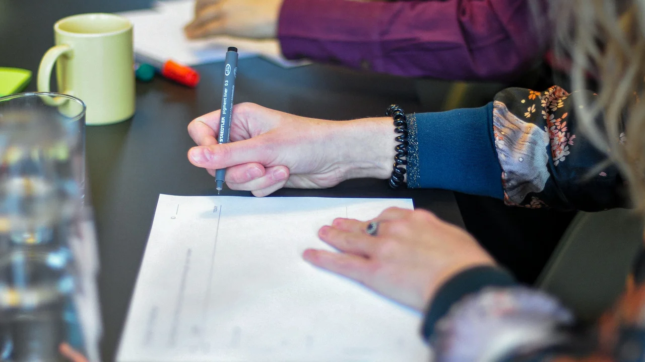 A close-up, high-angle shot focusing on a person's hands as they write on a white document with a thin black marker. The person wears a floral-patterned sleeve and a black coiled bracelet. The setting appears to be a collaborative workspace, with a yellow mug, blurred figures, and glassware on a dark table in the background.
