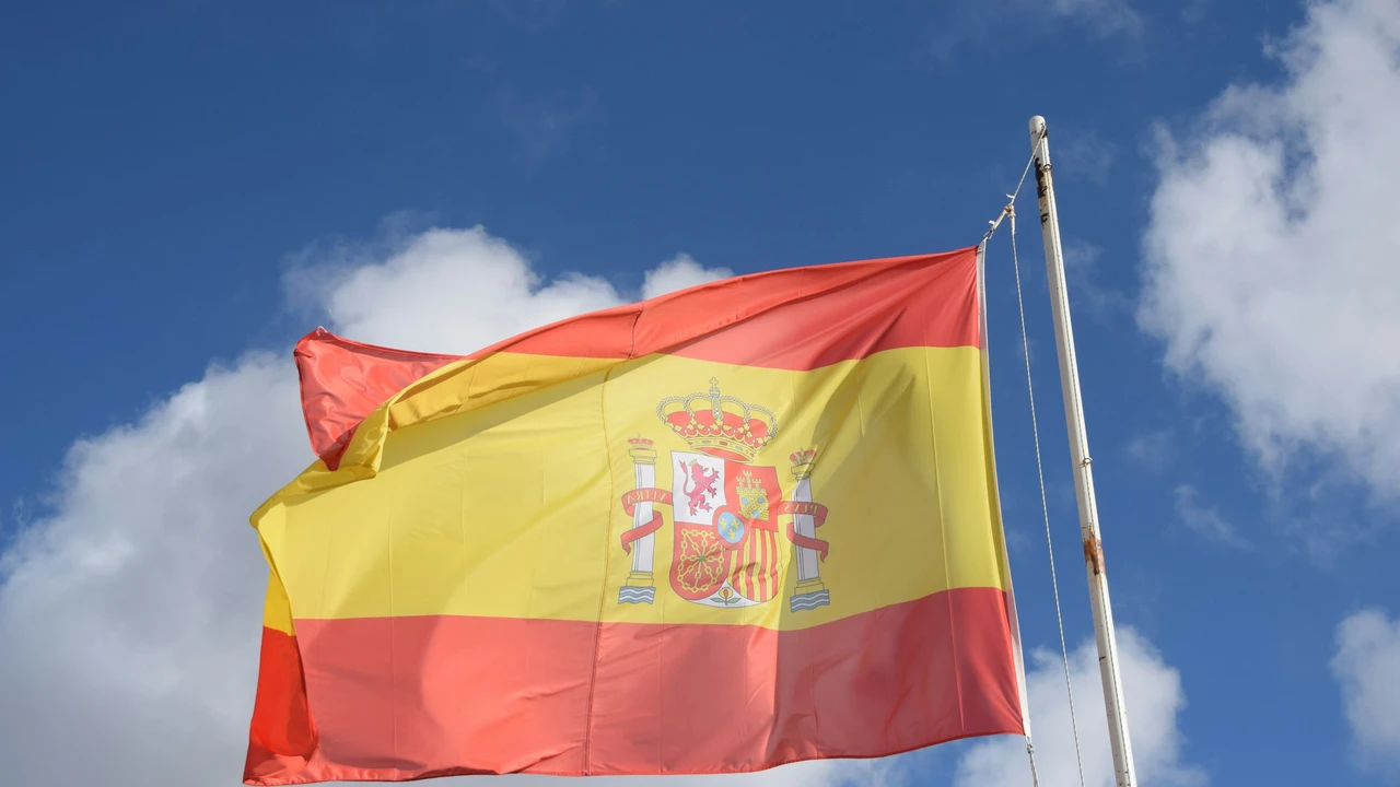 The national flag of Spain, featuring horizontal stripes of red and yellow with the royal coat of arms, flutters from a white flagpole against a bright blue sky with scattered white clouds.