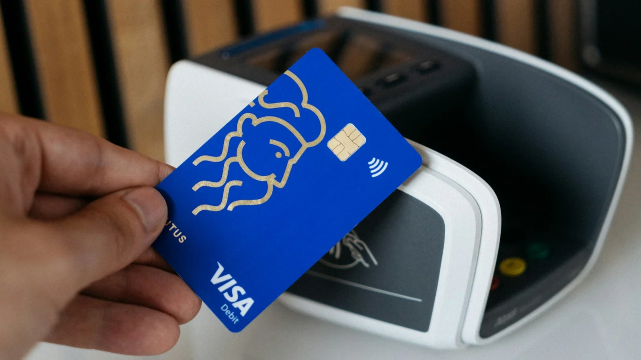 A close-up shot of a hand holding a blue Visa debit card near a white contactless payment terminal. The card features a gold stylized profile of a face. The background is slightly blurred, focusing on the action of preparing to make a payment.