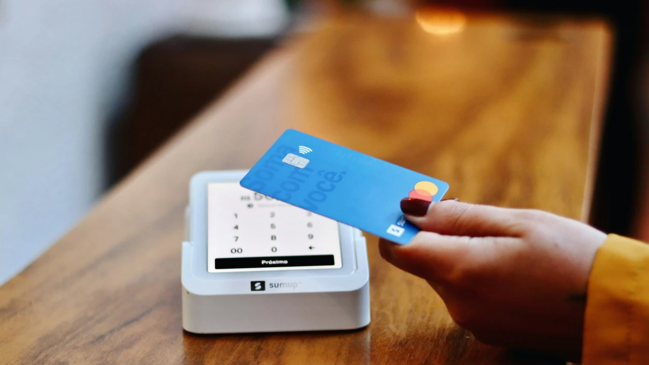 A close-up shot of a hand with maroon-painted nails holding a blue Mastercard toward a white 