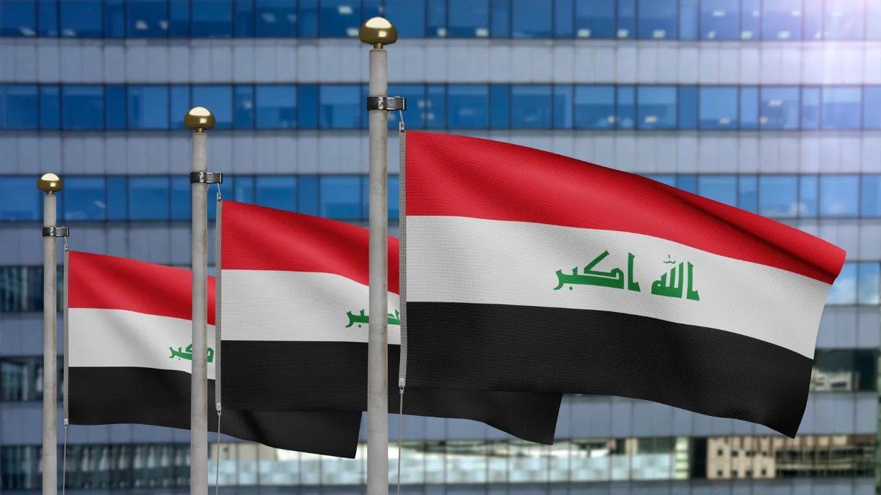 Iraqi national flags waving on flagpoles with modern glass office building background, red white black tricolor with green Arabic text “Allahu Akbar” visible in daylight.