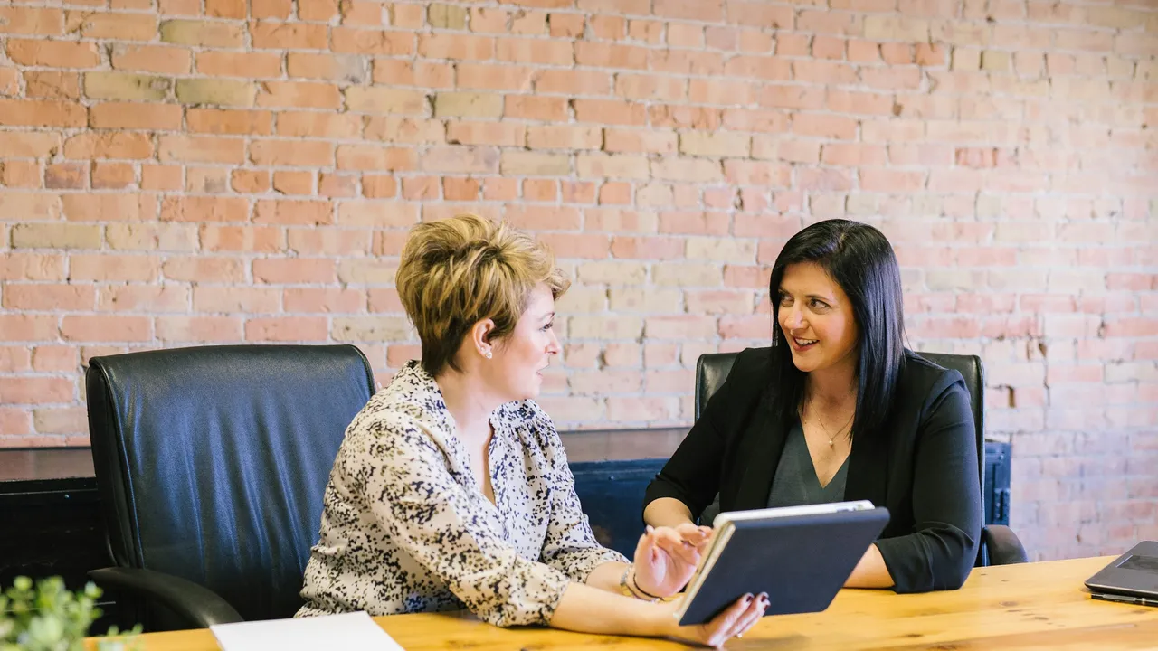 Two professional women sit at a wooden desk in front of a brick wall, engaged in a conversation while looking at a digital tablet.