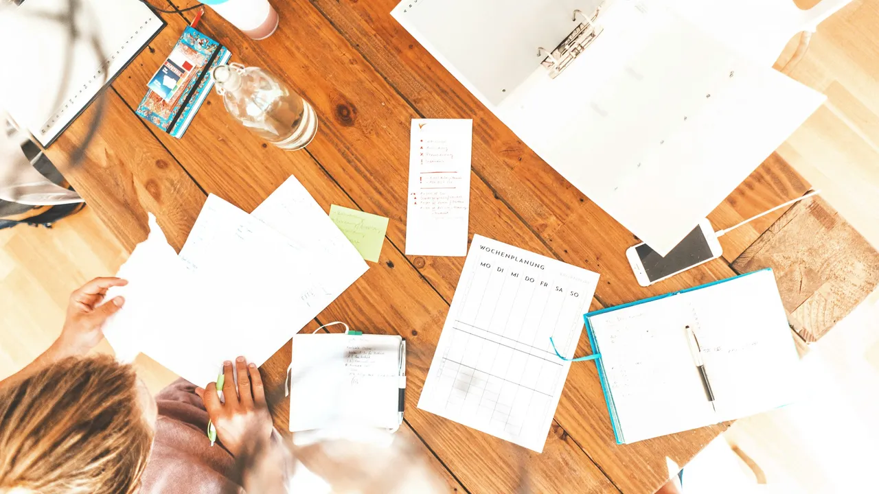 An overhead view of a person working at a rustic wooden table scattered with various organizational tools. The workspace includes a weekly planner, an open blue notebook with a pen, a white binder, a smartphone, and several loose papers being handled by the individual.