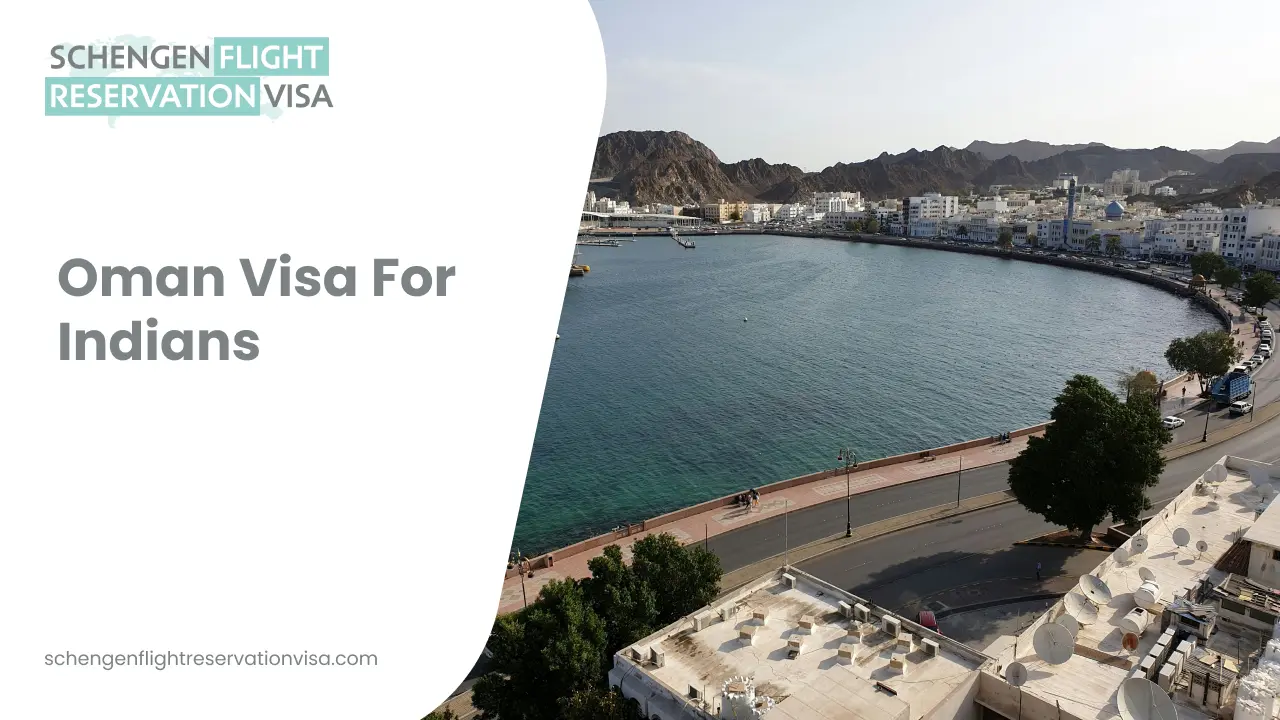 An aerial, wide-angle view of a coastal city in Oman, featuring a curved harbor with deep blue water. A paved promenade lined with trees and a roadway runs along the waterfront, while white, low-rise buildings and mosques are nestled at the base of rugged, brown mountains under a clear sky.