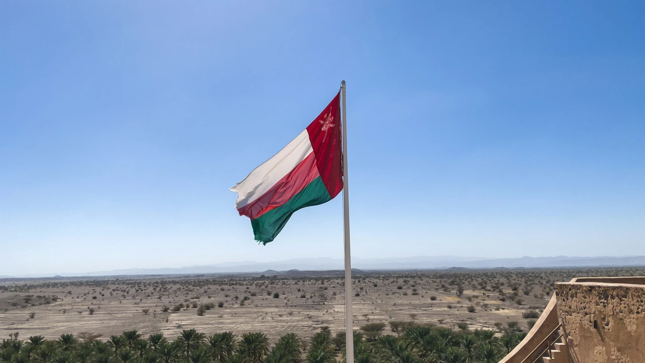 The national flag of Oman, featuring a vertical red stripe on the hoist side with the national emblem (the Khanjar) and three horizontal stripes of white, red, and green, waves from a flagpole. The flag is set against a vast, arid desert landscape dotted with sparse vegetation and palm trees under a bright blue sky.