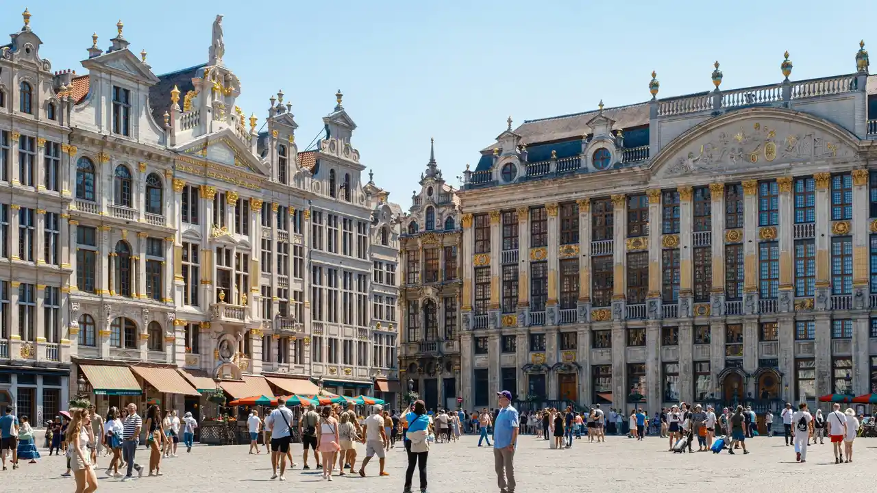  A crowded public square in Belgium, likely the Grand Place in Brussels, featuring ornate guildhalls with gold detailing and classical architecture under a clear blue sky.