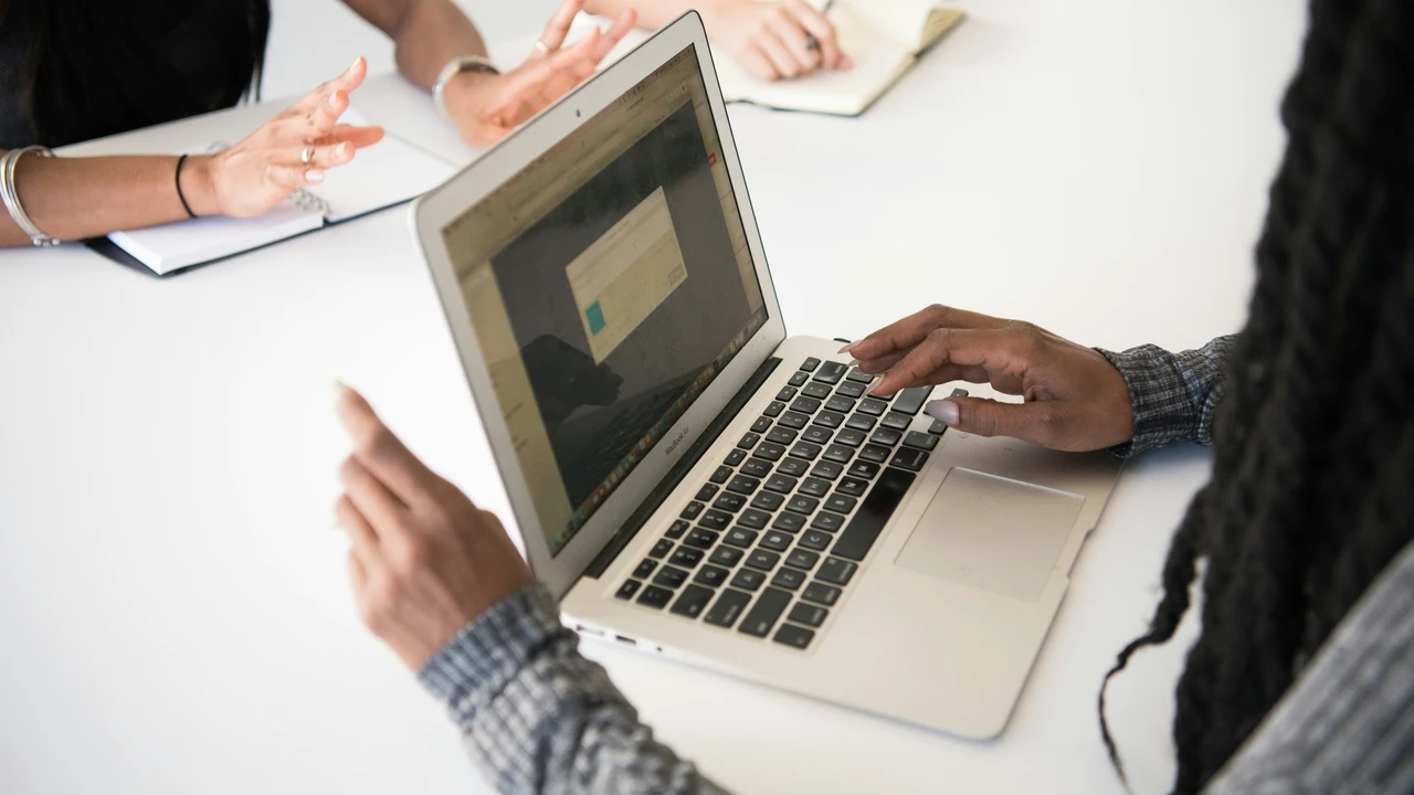An overhead view of a person with long braids typing on a silver laptop at a white table. Across from them, another person’s hands are visible, gesturing while they talk, with a notebook open nearby. The scene suggests a professional discussion or a consultation meeting.