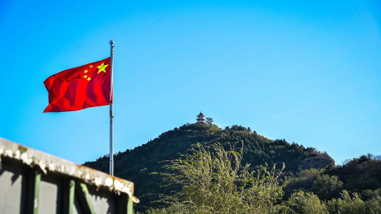 A red Chinese national flag fluttering on a flagpole in the foreground, with a traditional multi-tiered pagoda situated atop a lush green hill under a clear blue sky in the background.
