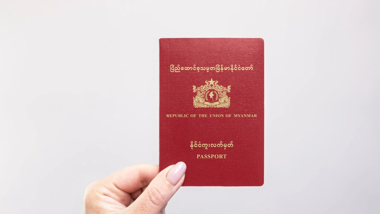 Close-up of a hand holding a red Republic of the Union of Myanmar passport with gold emblem and Burmese script on the cover against a light gray background.