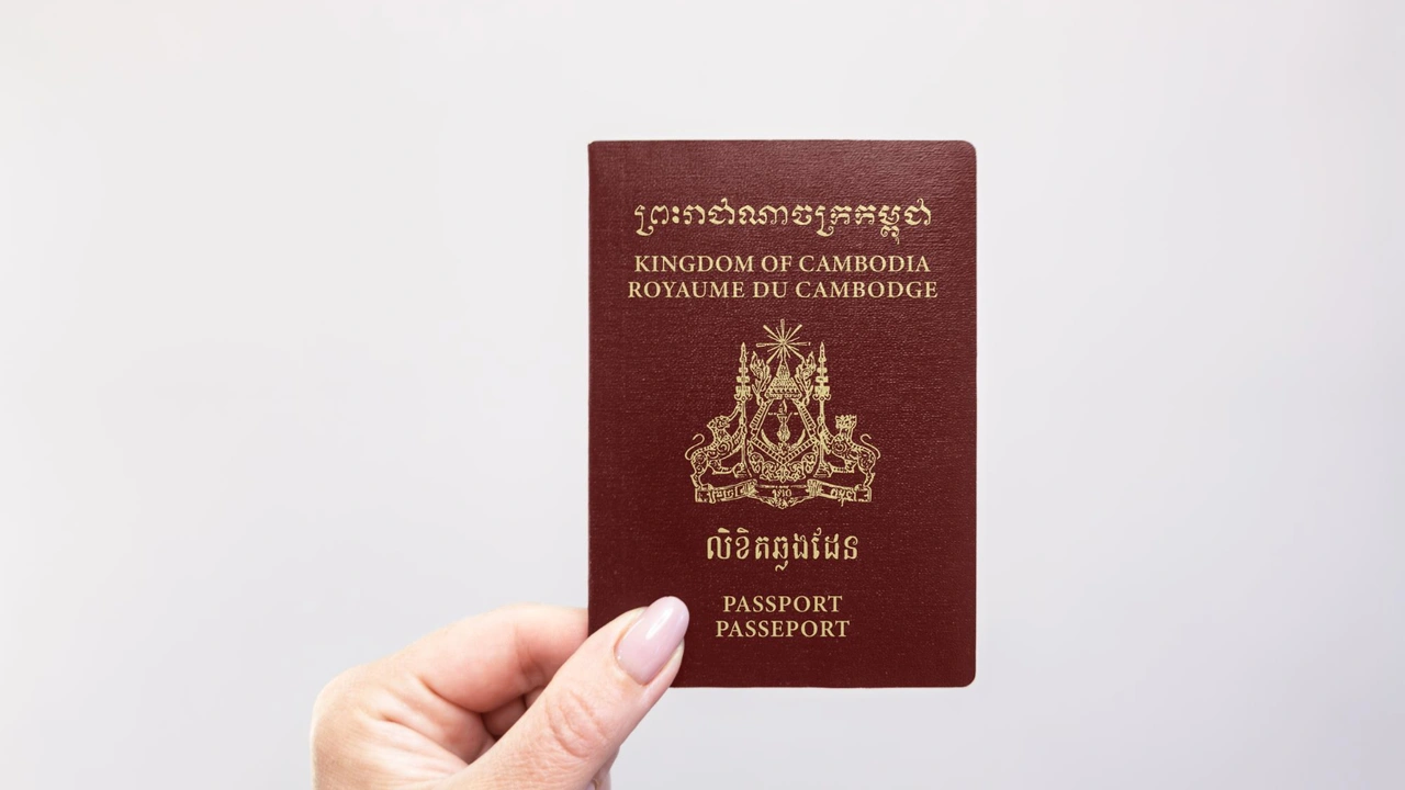 Close-up of a hand holding a Cambodia passport with gold emblem and “Kingdom of Cambodia” text on a brown cover, isolated on a light background, symbolizing international travel and citizenship documentation.