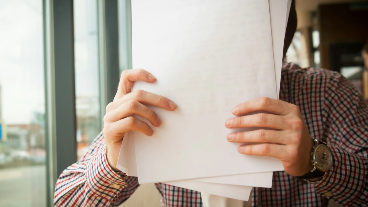 A close-up shot of a person's hands holding a thick stack of white, blank papers in front of their chest. The person is wearing a red and blue checkered shirt and a silver wristwatch. The background is slightly blurred, showing a window with bright daylight.