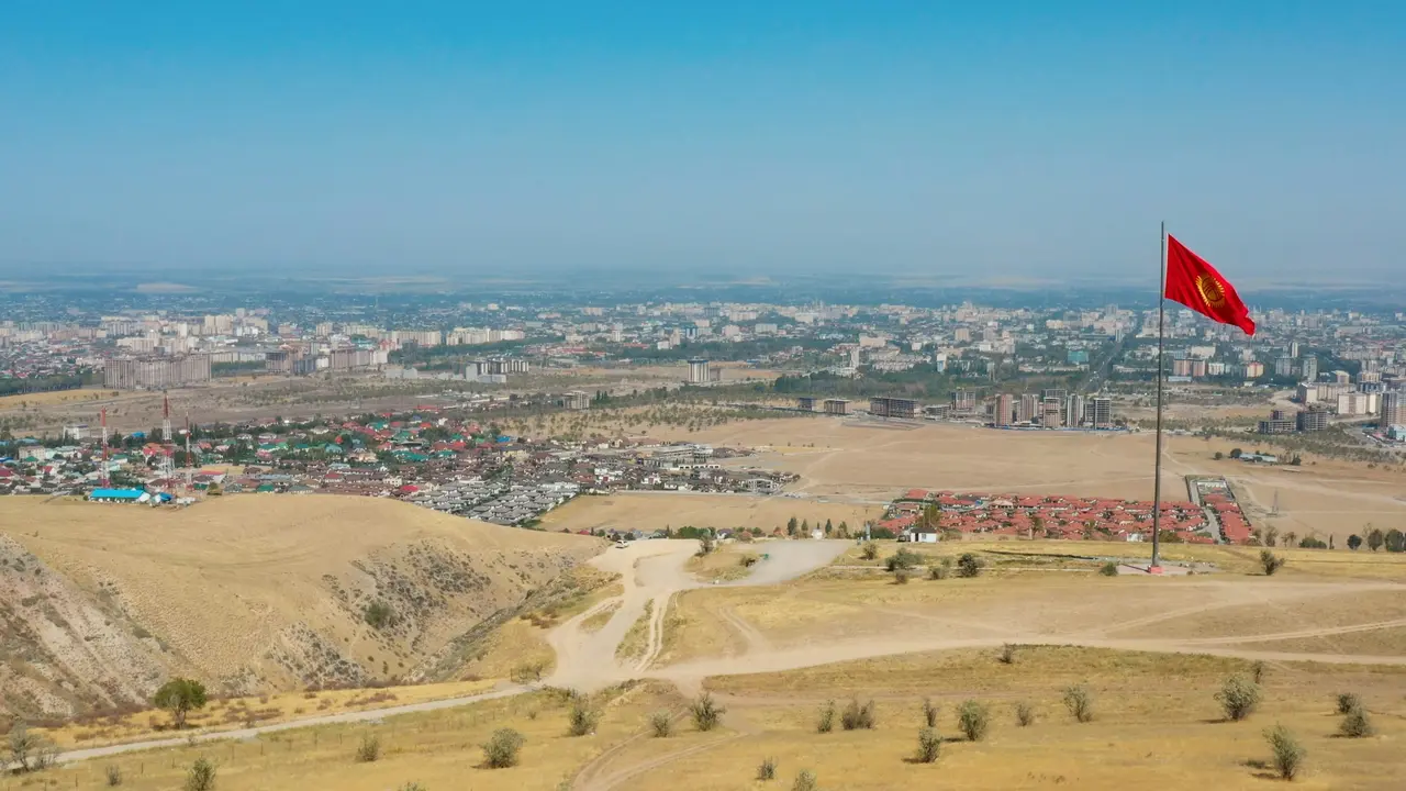 An elevated, wide-angle view of a city in Kyrgyzstan under a bright blue sky. In the foreground, a large red national flag flies on a tall pole atop a dry, grassy hill overlooking the urban sprawl.