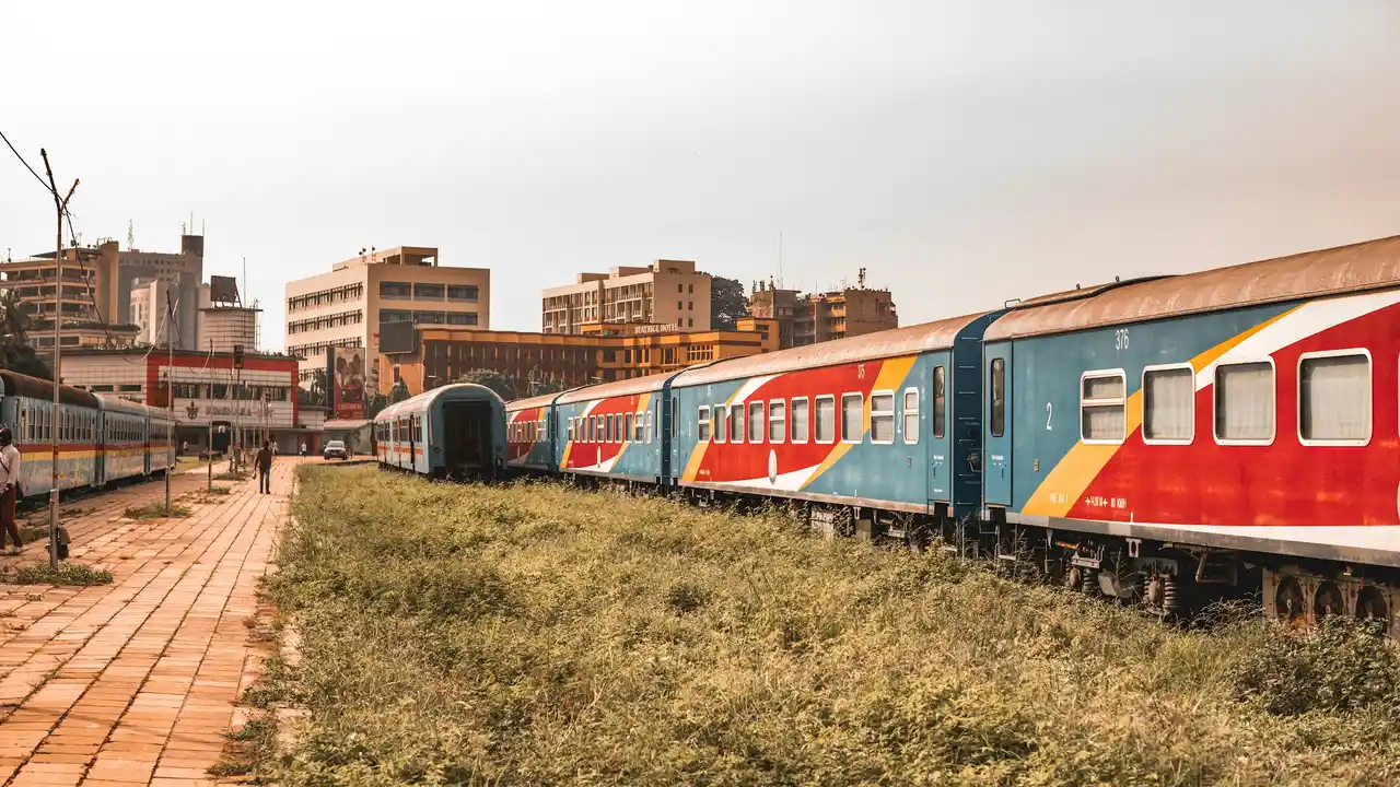 Colorful passenger train carriages standing on railway tracks at an urban train station, with city buildings in the background and grass growing alongside the platform.
