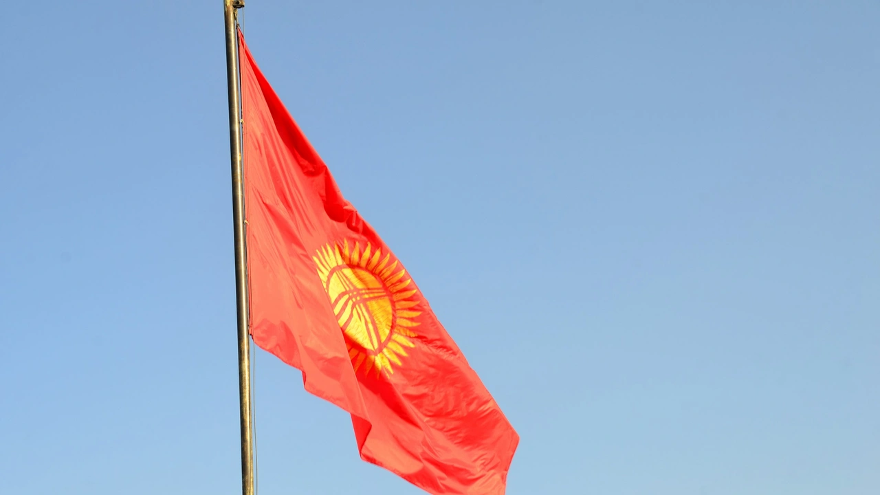 A close-up photograph of the Kyrgyzstan national flag—a red field with a yellow sun in the center—waving from a flagpole against a clear, bright blue sky.