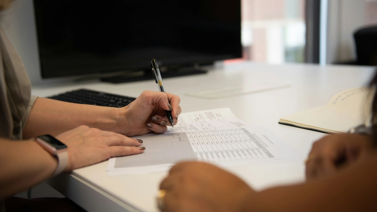 An over-the-shoulder view of a person wearing a smartwatch signing a detailed application form or document with a black pen on a white desk. A computer monitor and a notebook are visible in the background, representing the administrative process of waiting for visa processing.