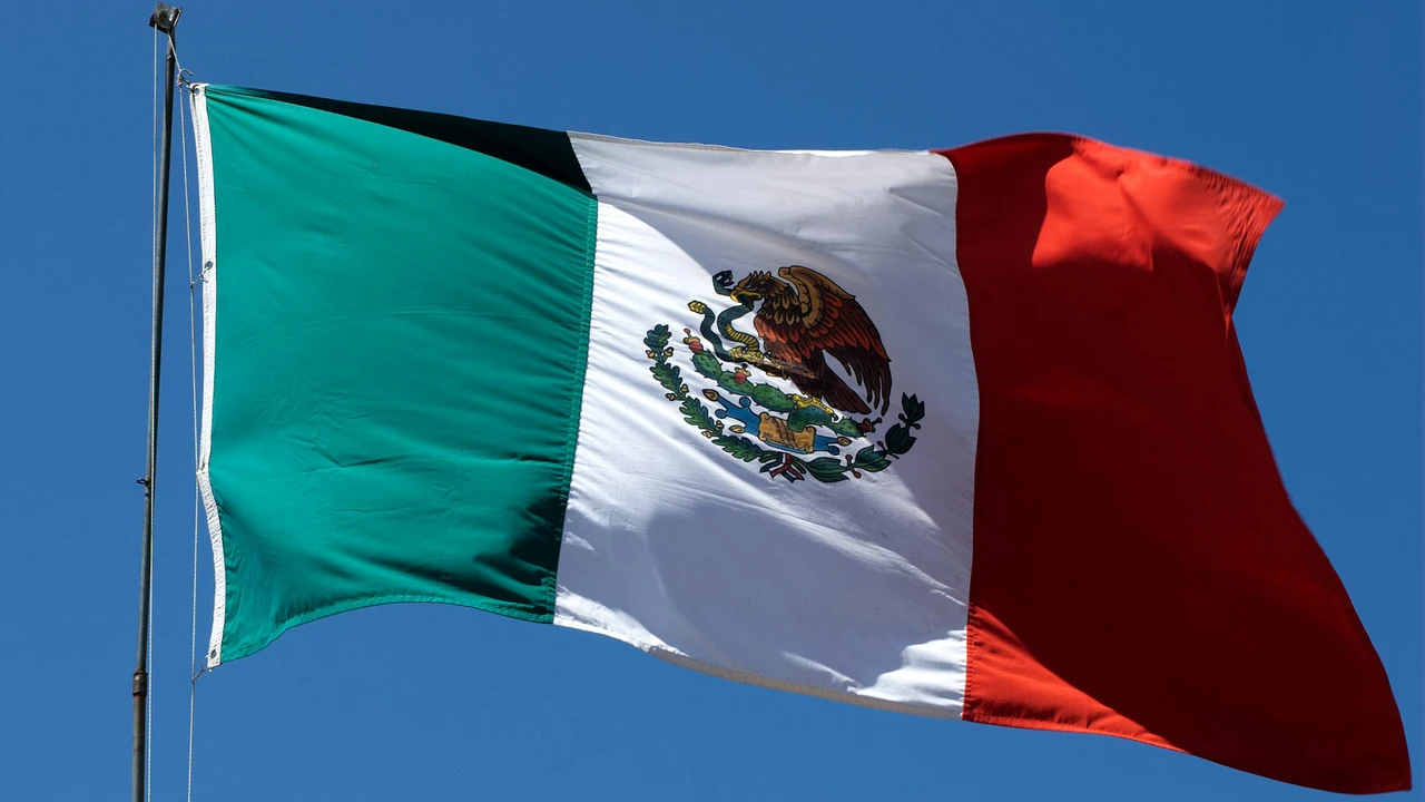 A close-up photograph of the Mexican flag waving prominently against a clear, solid blue sky. The flag features its characteristic vertical stripes of green, white, and red, with the national