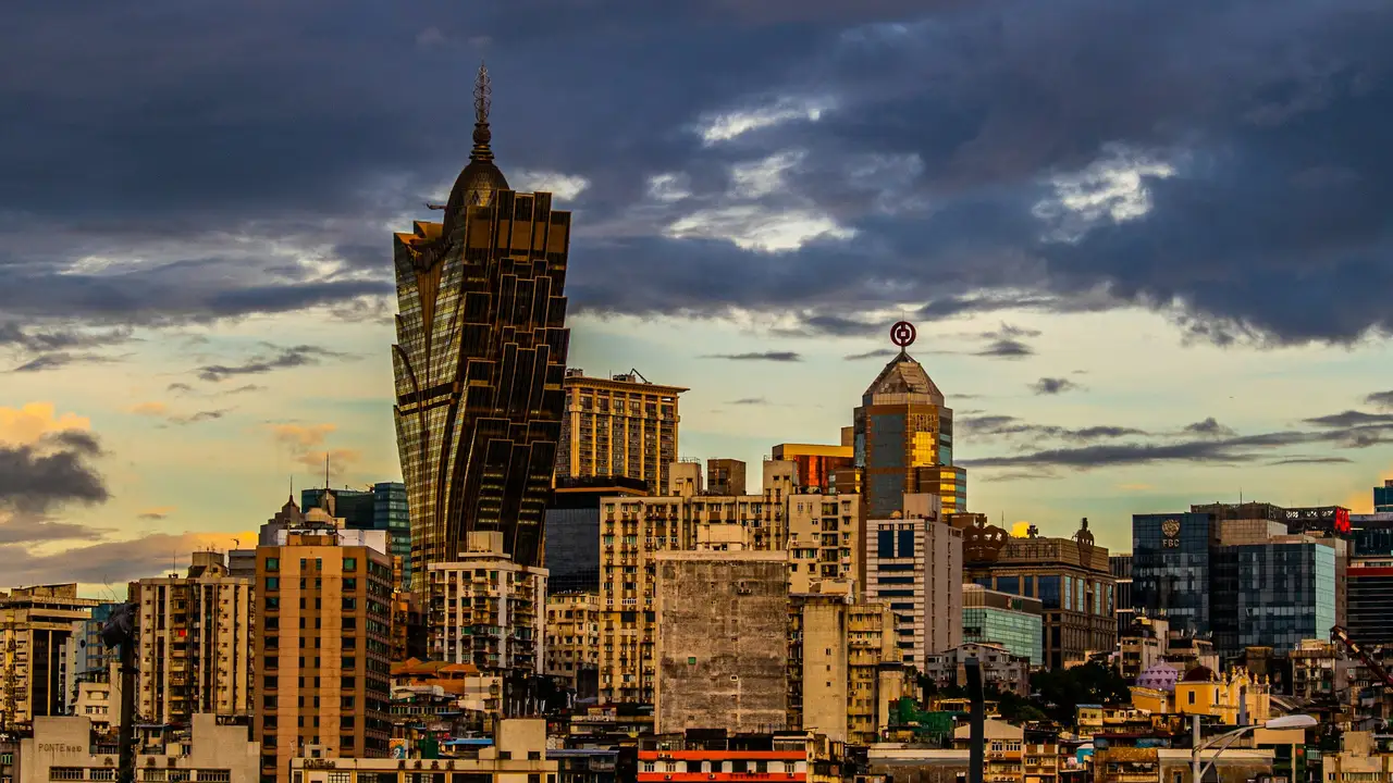 Alt text for the final image provided is as follows: A vibrant twilight cityscape of Macau, featuring the iconic gold, lotus-flower-shaped Grand Lisboa hotel and casino as the central landmark. The skyline shows a dense mix of modern high-rises and older urban buildings under a soft, darkening sky. On the left, a white graphic overlay includes the text