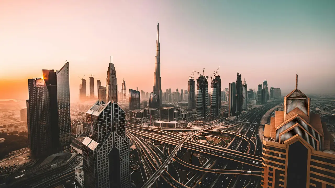 An aerial perspective of the Dubai skyline at sunset, dominated by the Burj Khalifa, the world's tallest building. In the foreground, a complex network of multi-level highway interchanges on Sheikh Zayed Road is filled with moving traffic
