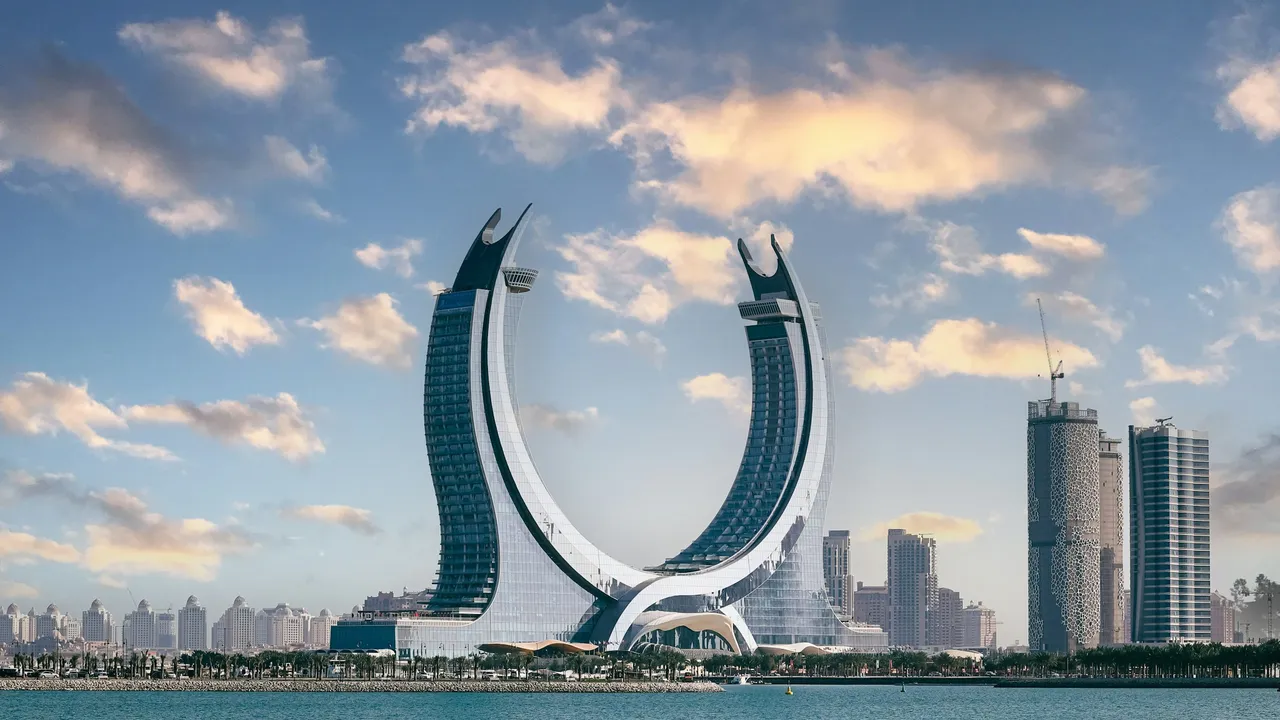 The Katara Towers, a striking crescent-shaped architectural landmark in Lusail, Qatar, stand prominently against a soft sunset sky with scattered clouds. The twin curved towers face each other over a waterfront, with the Doha skyline and various high-rise buildings visible in the background across the blue water