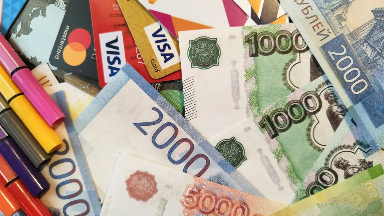 A close-up, top-down view of a cluttered desk featuring several 2000 and 1000 denomination banknotes mixed with various credit and debit cards, including Visa and Mastercard. A row of colorful pens in shades of pink, yellow, and blue is visible on the left side of the frame