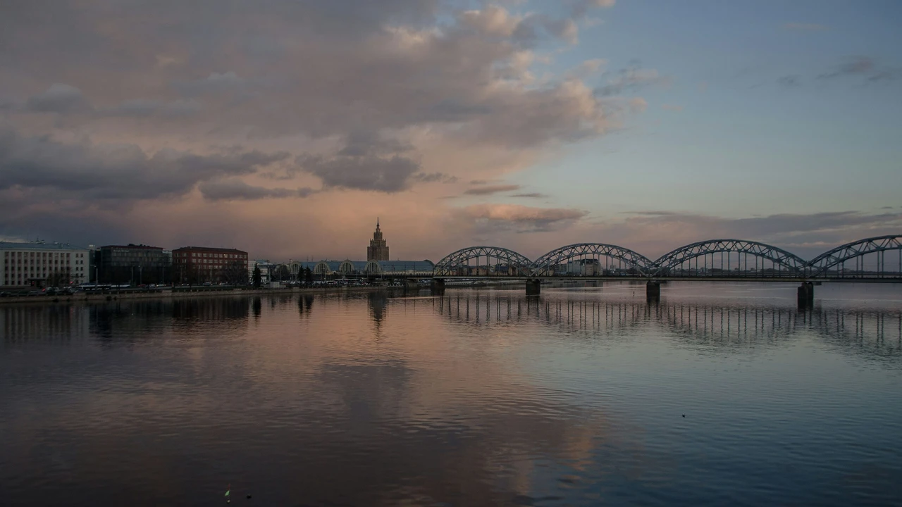 A wide-angle landscape photo of the Daugava River in Riga, Latvia, at sunset. A long, arched iron railway bridge crosses the calm water, which reflects the soft pink and purple hues of the evening sky. In the background, the city skyline features the silhouette of the Latvian Academy of Sciences building under a blanket of heavy clouds.