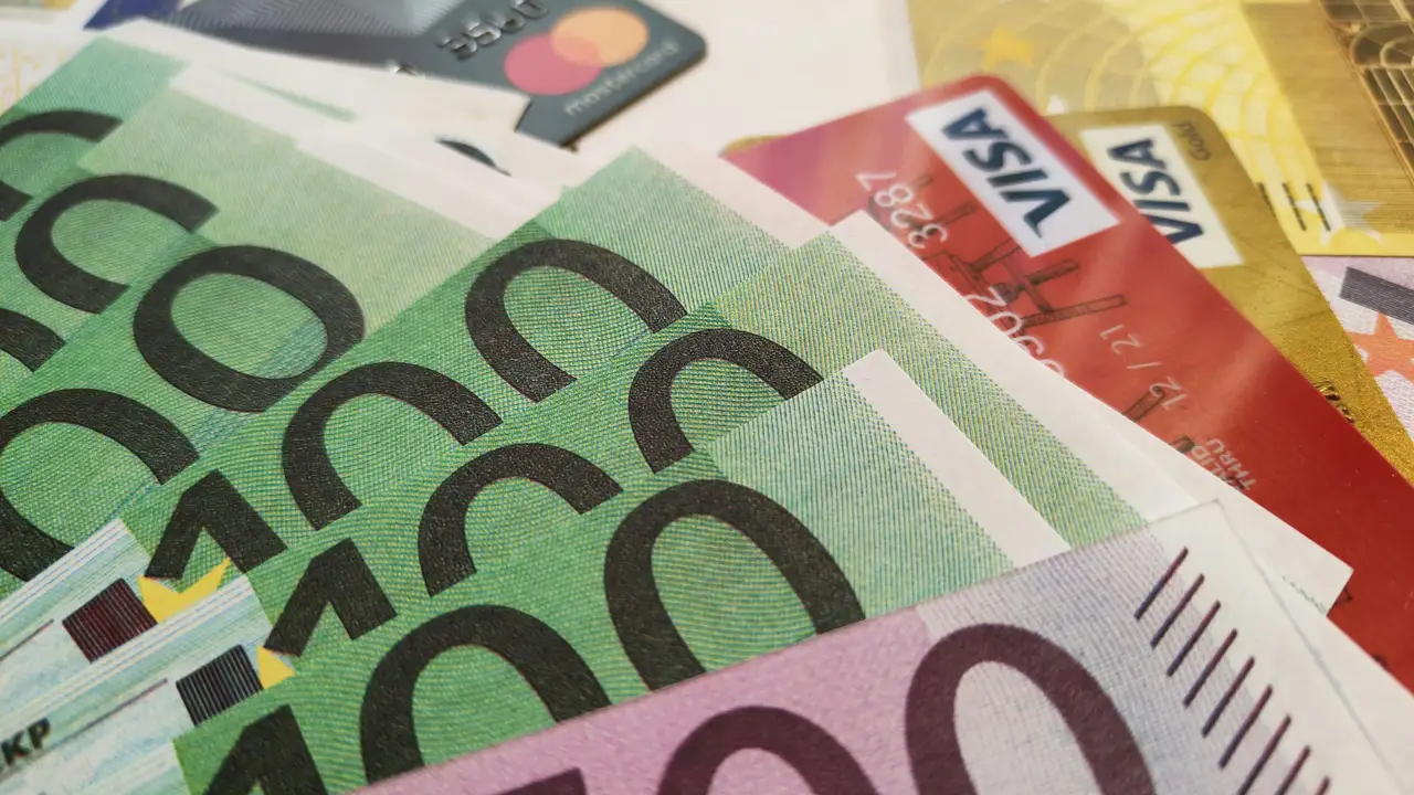 A high-angle, close-up shot of several Euro banknotes—including green 100 Euro notes and purple 500 Euro notes—intermingled with various credit and debit cards, including visible Visa and Mastercard logos. The currency and cards are spread out in a layered pile on a flat surface.