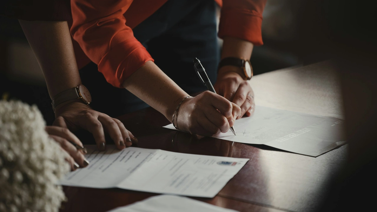 A close-up, high-angle shot focusing on the hands of two people at a wooden desk, engaged in signing and reviewing official documents. One person, wearing an orange long-sleeved shirt and a watch, is actively signing a paper with a black pen, while the other person's hand rests on a separate document nearby. The scene is warmly lit, emphasizing a professional or legal atmosphere.