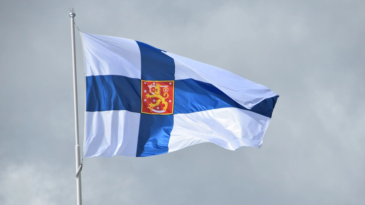 Flag of Finland waving on a flagpole against a cloudy sky, featuring a blue Nordic cross on a white background with the Finnish coat of arms in the center.