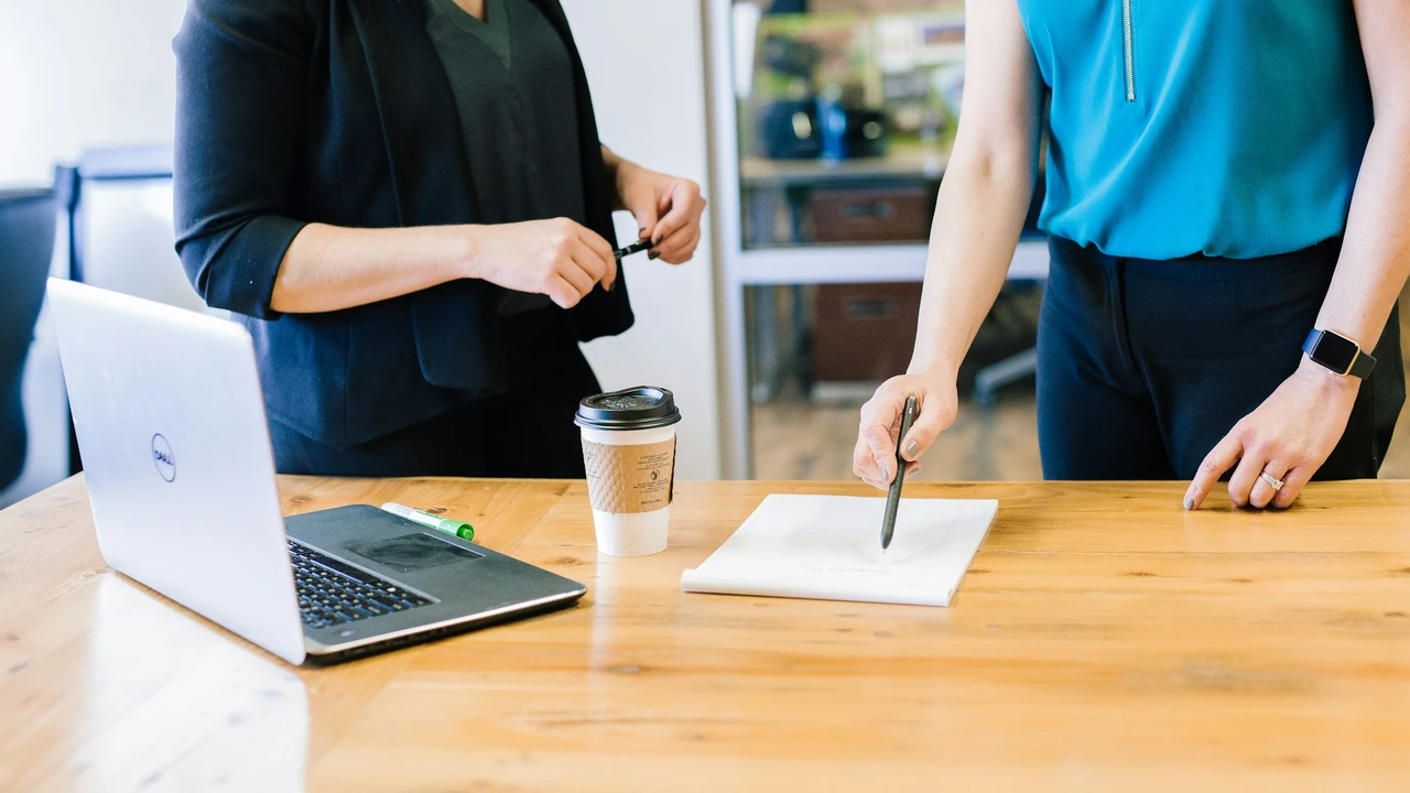 An office setting featuring two people standing at a light-colored wooden desk during a collaborative discussion. One person, wearing a bright teal sleeveless top and dark trousers, is pointing a pen toward a white notepad on the table. The other person, dressed in a black blazer, stands opposite them. A silver laptop and a disposable coffee cup are also on the desk, creating a professional and focused atmosphere.