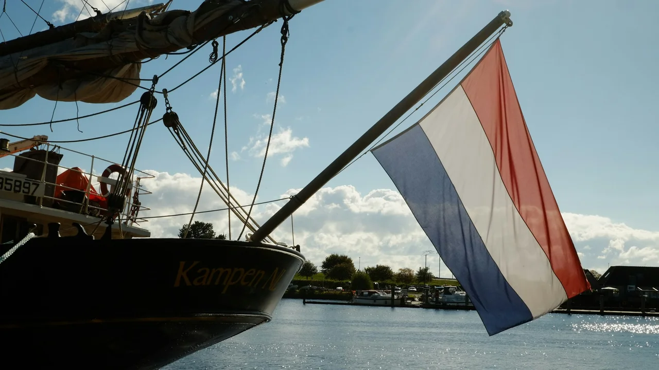 A low-angle shot of the Dutch national flag (red, white, and blue horizontal stripes) flying from the stern of a traditional wooden sailing vessel named