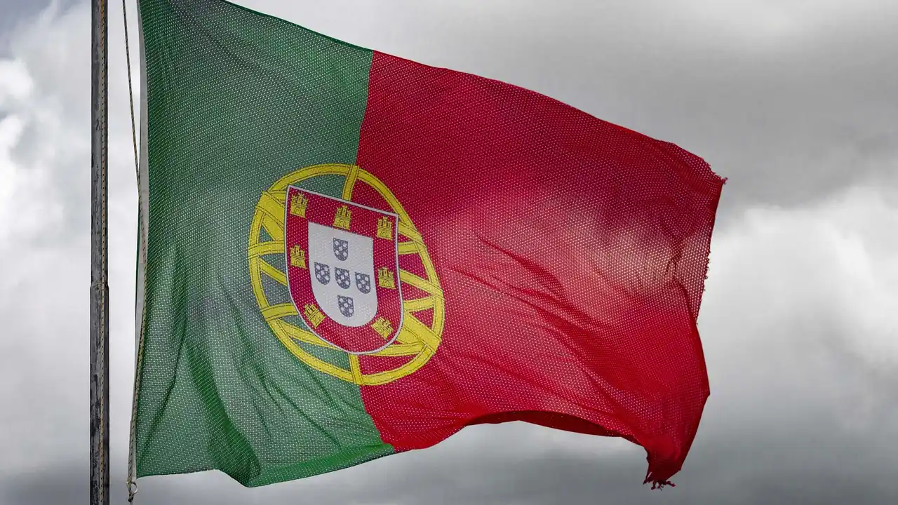 A close-up view of the national flag of Portugal, featuring its vertical green and red bands and the national coat of arms, waving against a backdrop of grey, overcast clouds.