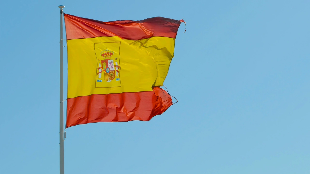 Spanish national flag with red and yellow horizontal stripes and the coat of arms, waving on a flagpole against a clear blue sky.