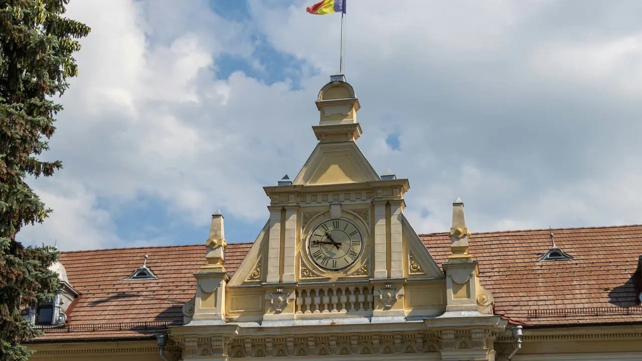 The Romanian national flag, with its vertical stripes of blue, yellow, and red, flies from a flagpole atop a classic yellow building. The structure features a prominent central pediment housing a large analog clock with Roman numerals, set against a background of a brown tiled roof and a blue sky with soft white clouds