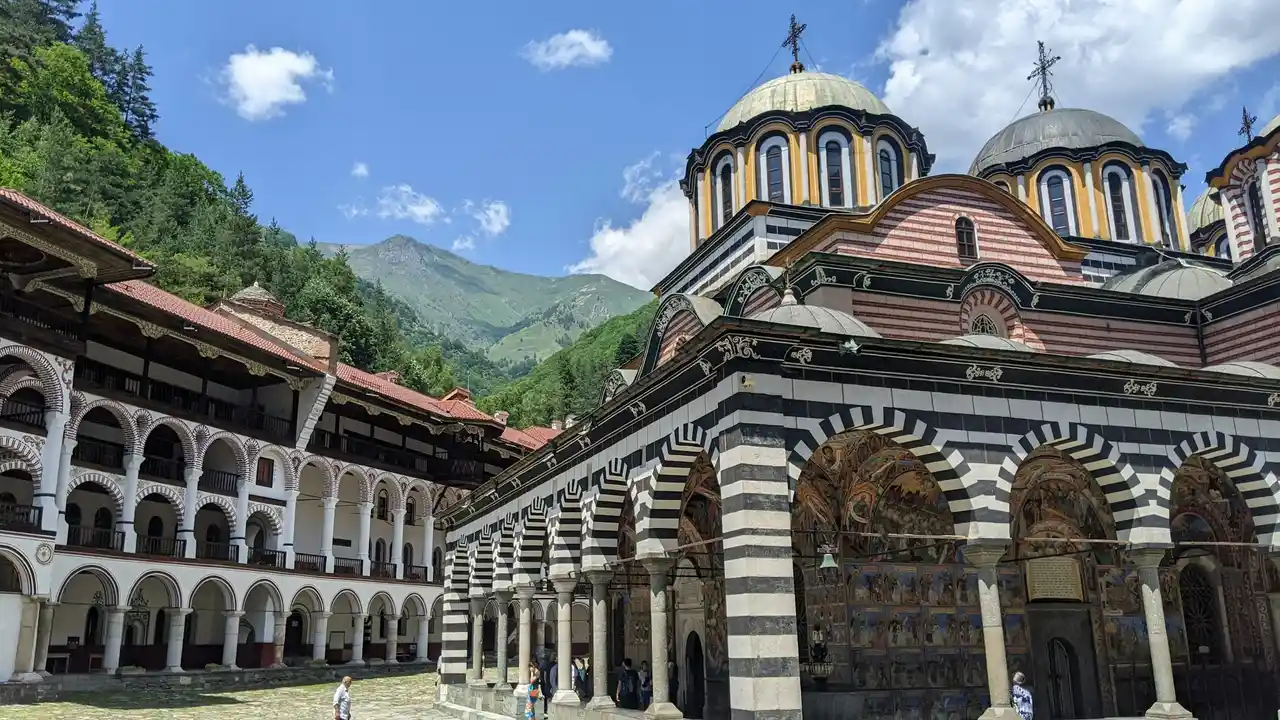 Rila Monastery in Bulgaria with striped arches, domed church, mountain backdrop, and historic Orthodox architecture under blue sky