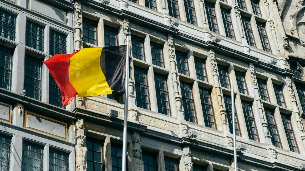 A Belgian flag—featuring vertical stripes of black, yellow, and red—waves in front of a grand, historic building with ornate stone carvings and numerous tall, multi-paned windows.