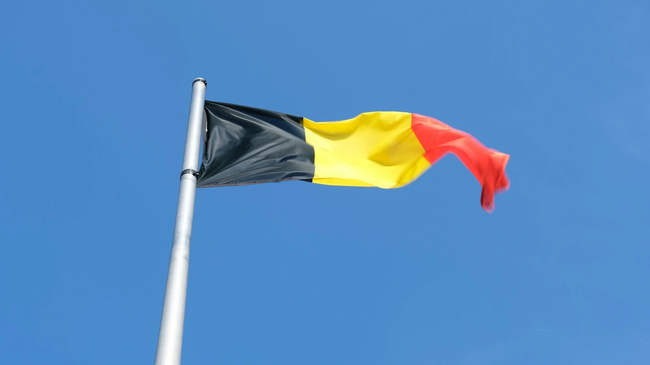 The national flag of Belgium, featuring vertical stripes of black, yellow, and red, waving from a tall silver flagpole against a clear, bright blue sky