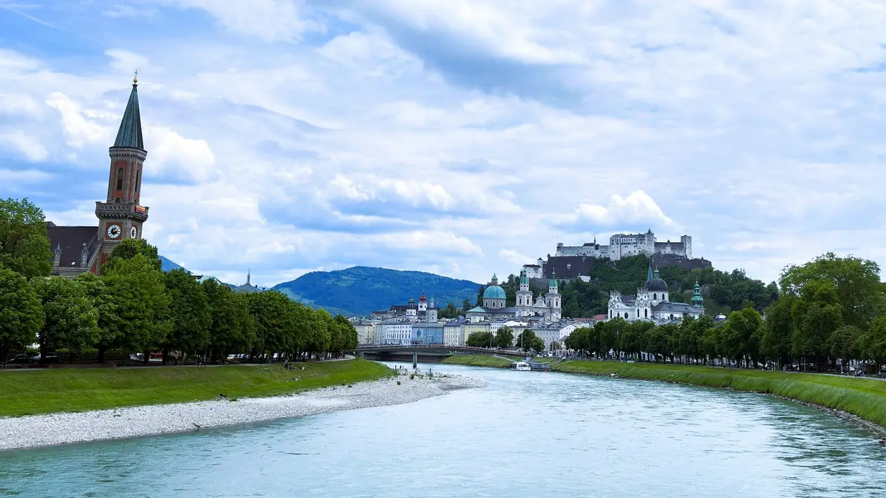 A wide-angle landscape view of the city of Salzburg, Austria, featuring a calm river flowing through the center with a gravel bank on one side. Green trees line both sides of the river, and historic buildings with various domes and steeples are visible in the background. Hohensalzburg Fortress sits atop a hill overlooking the city under a cloudy blue sky.