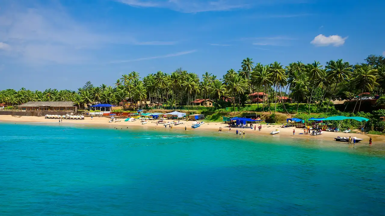 A vibrant, sunny view of a tropical beach in Goa, featuring bright turquoise water, a sandy shoreline with colorful beach shacks and boats, and a lush backdrop of tall palm trees under a clear blue sky.