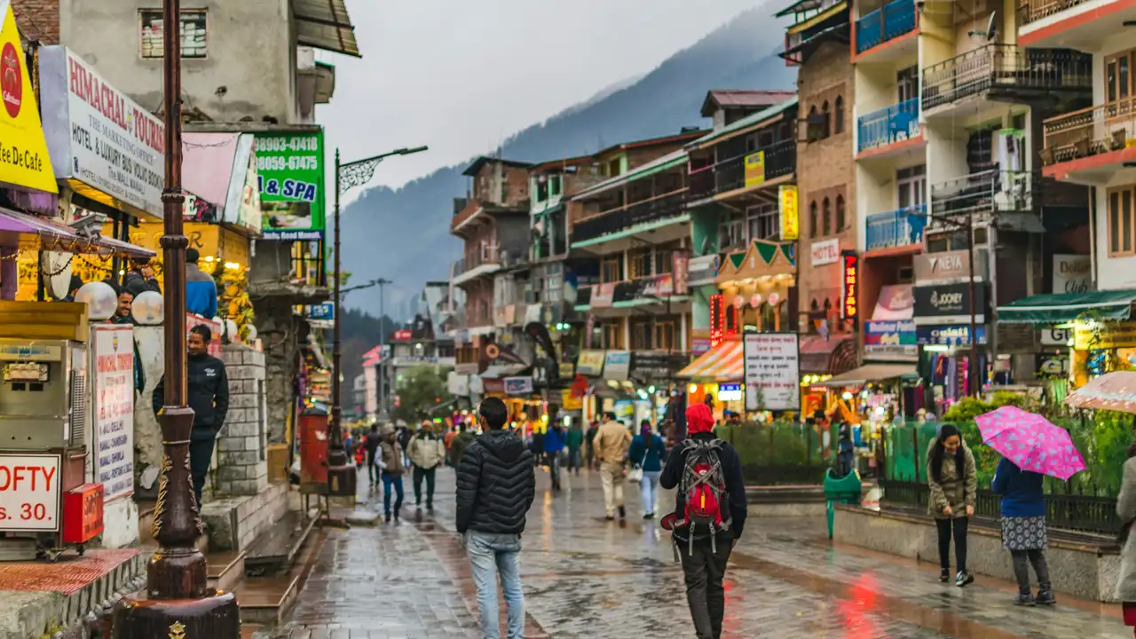 A bustling, wet street scene at Mall Road in Manali, India, featuring pedestrians walking past colorful multi-story buildings and storefronts under an overcast sky with misty mountains in the distance.