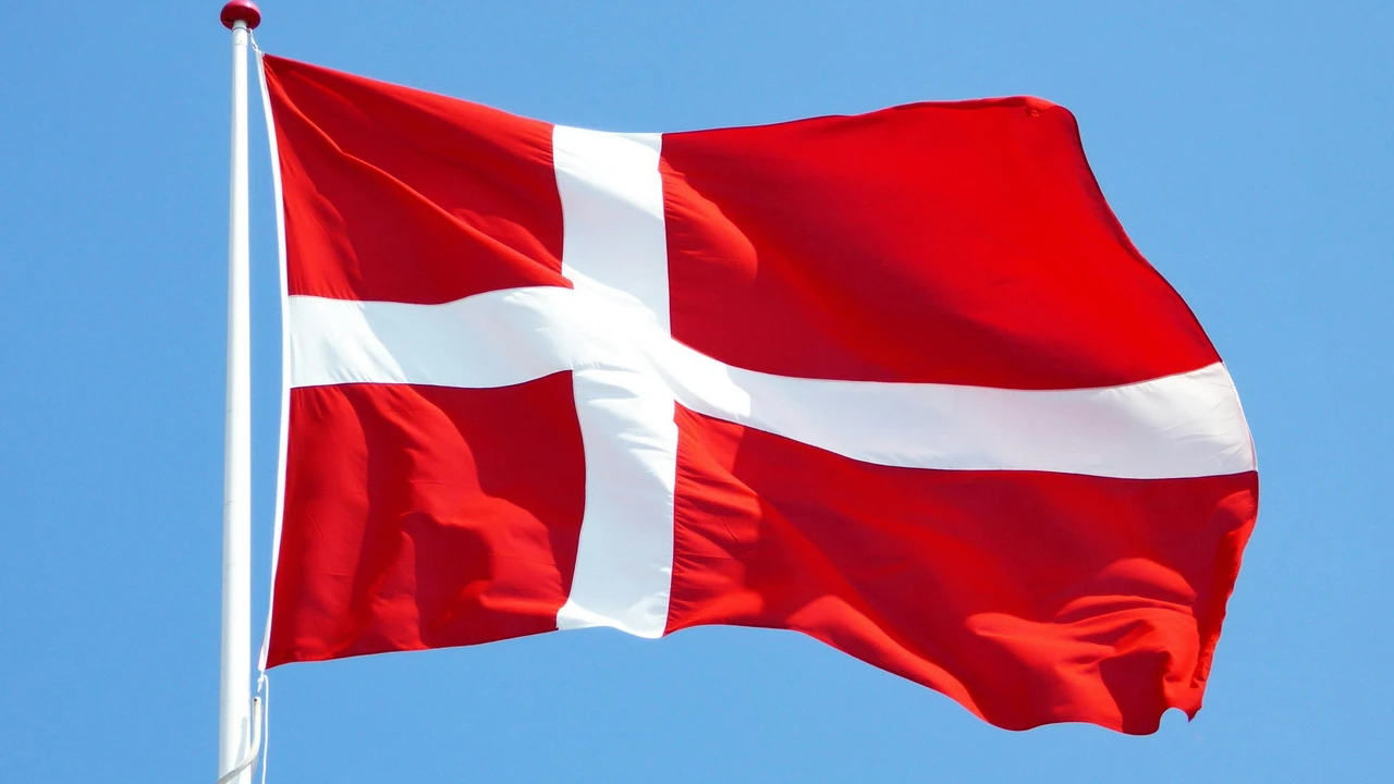 The national flag of Denmark, a red flag with a white Nordic cross, waving on a flagpole against a clear blue sky.